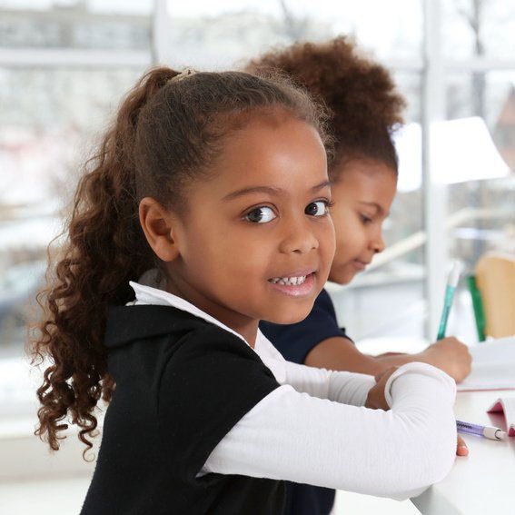 Two young girls, both with dark skin, seated at a desk, looking at the camera. The girl in front is smiling.