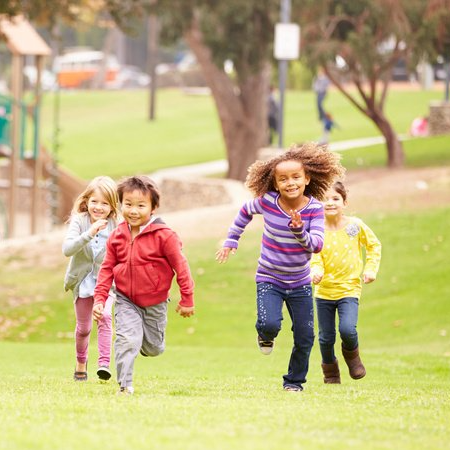 Children of various ethnicities running on a grassy park. They are smiling and appear to be playing. Trees and a playground are in the background.
