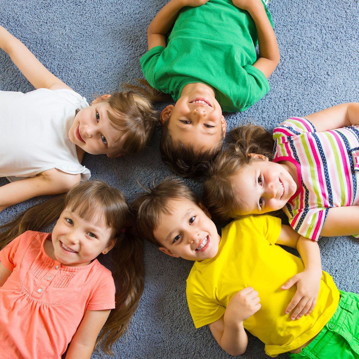 Six smiling children lying on a blue carpet, forming a circle with their heads together.
