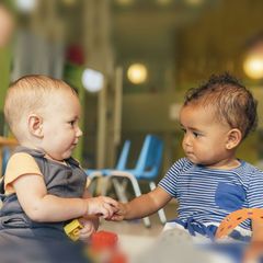 Two babies, one fair-skinned, one mixed-race, sit facing each other in a play area, holding hands.