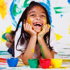 A smiling child covered in paint rests their chin on hands, in front of colorful paint splotches and paint bowls.
