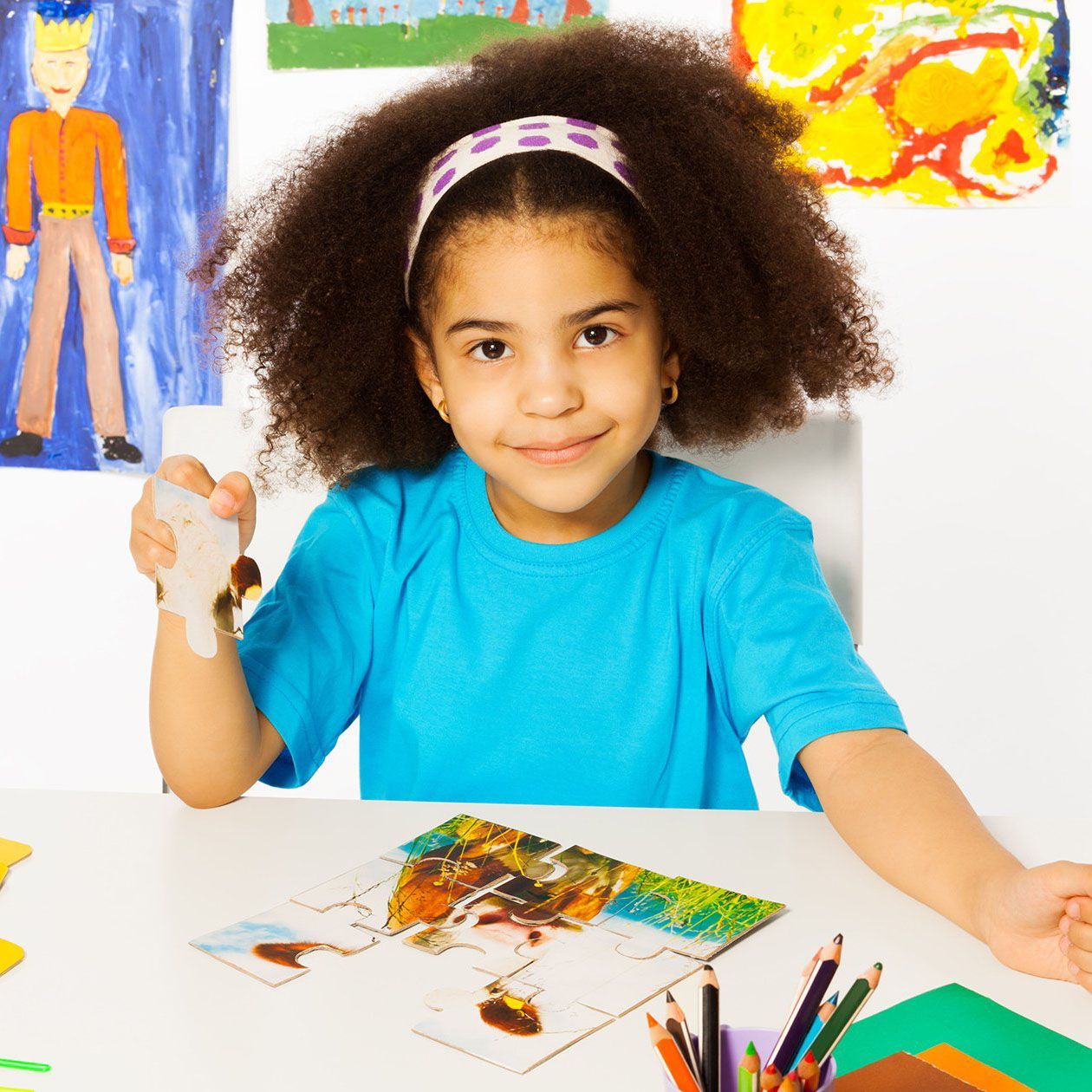 Young girl with curly hair wearing a blue shirt and a headband smiles while holding a paintbrush, seated at a table with artwork.