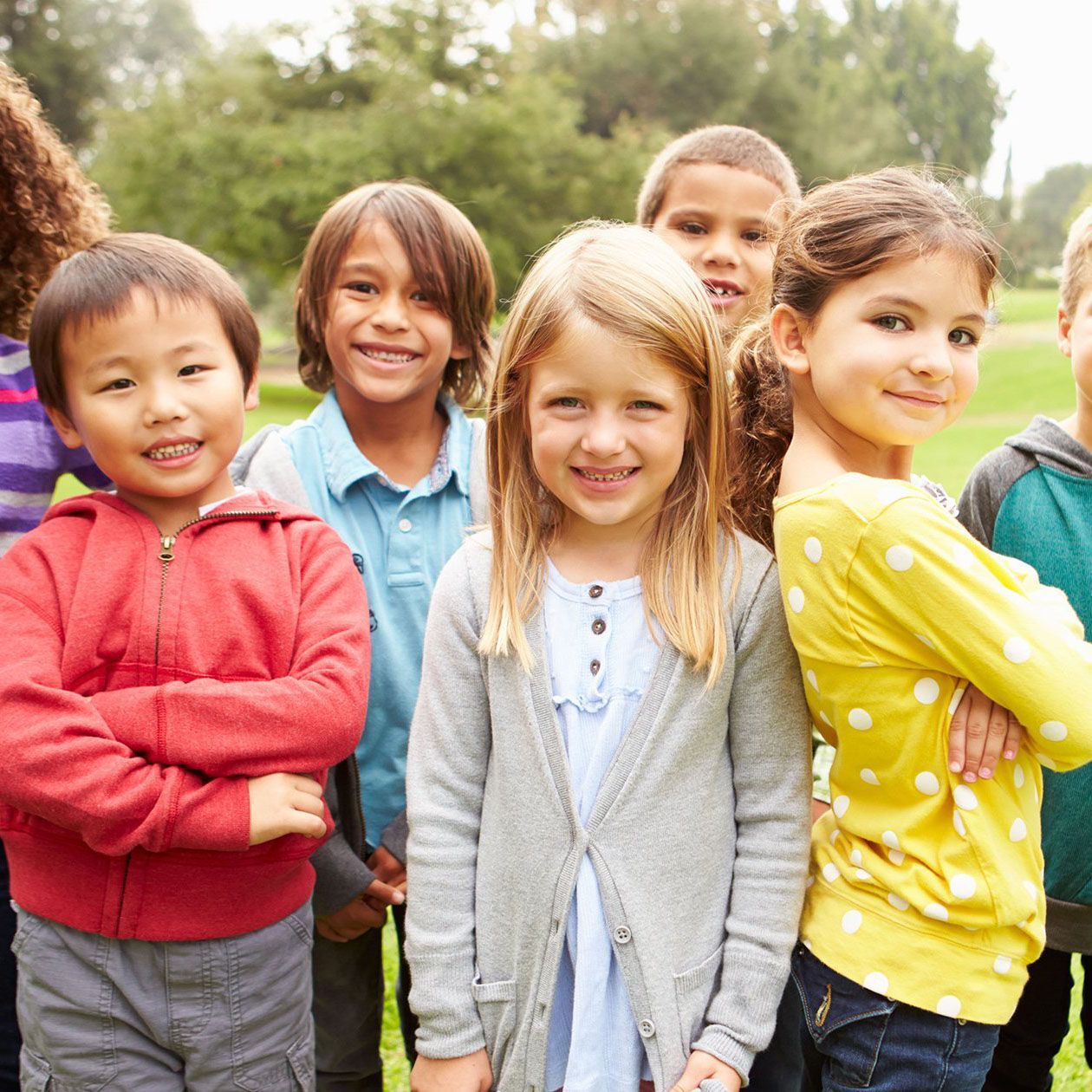 Group of smiling children of diverse ethnicities standing outdoors. They are in casual clothes, with a grassy background.