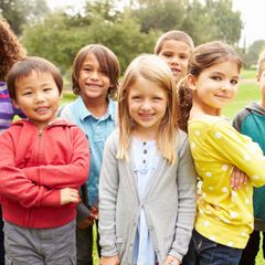 Group of smiling children of diverse ethnicities standing outdoors. They are in casual clothes, with a grassy background.