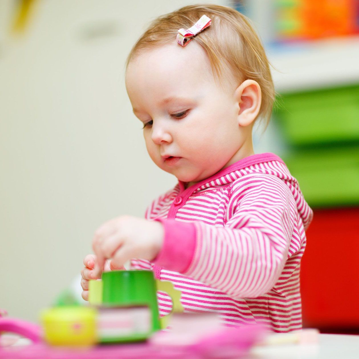 A toddler with a pink striped hoodie and hair clip plays with green and pink toys at a table.