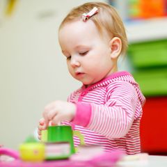 A toddler with a pink striped hoodie and hair clip plays with green and pink toys at a table.
