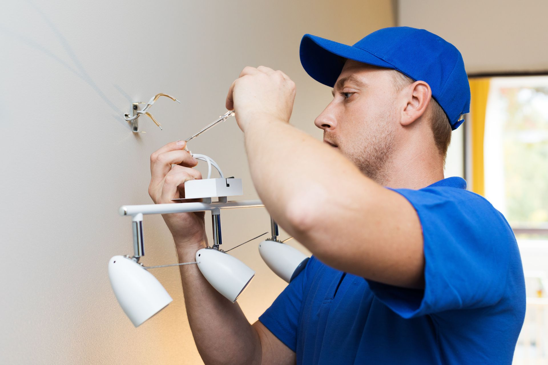 Electrician in blue cap and shirt installing a ceiling light fixture indoors