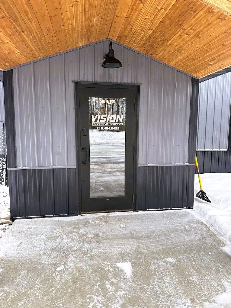 Small covered entrance with black door, gray siding, wood ceiling, and snow-covered concrete floor.