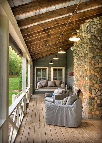 Covered wooden porch with cushioned chairs, hanging lights, and a stone wall overlooking a grassy yard