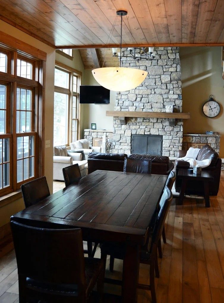 Dining room with dark wood table, stone fireplace, large windows, and warm wooden ceiling