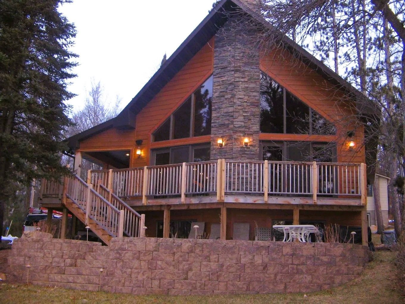 Wooden cabin with stone chimney and wraparound porch in a wooded setting at dusk