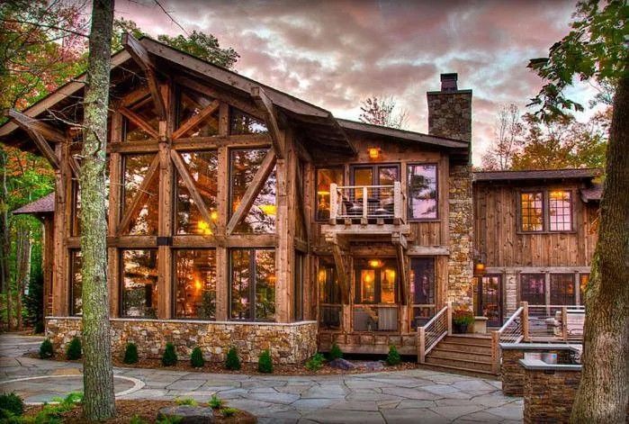 Rustic wooden house with large windows and a stone walkway, lit warmly at dusk under a cloudy sky
