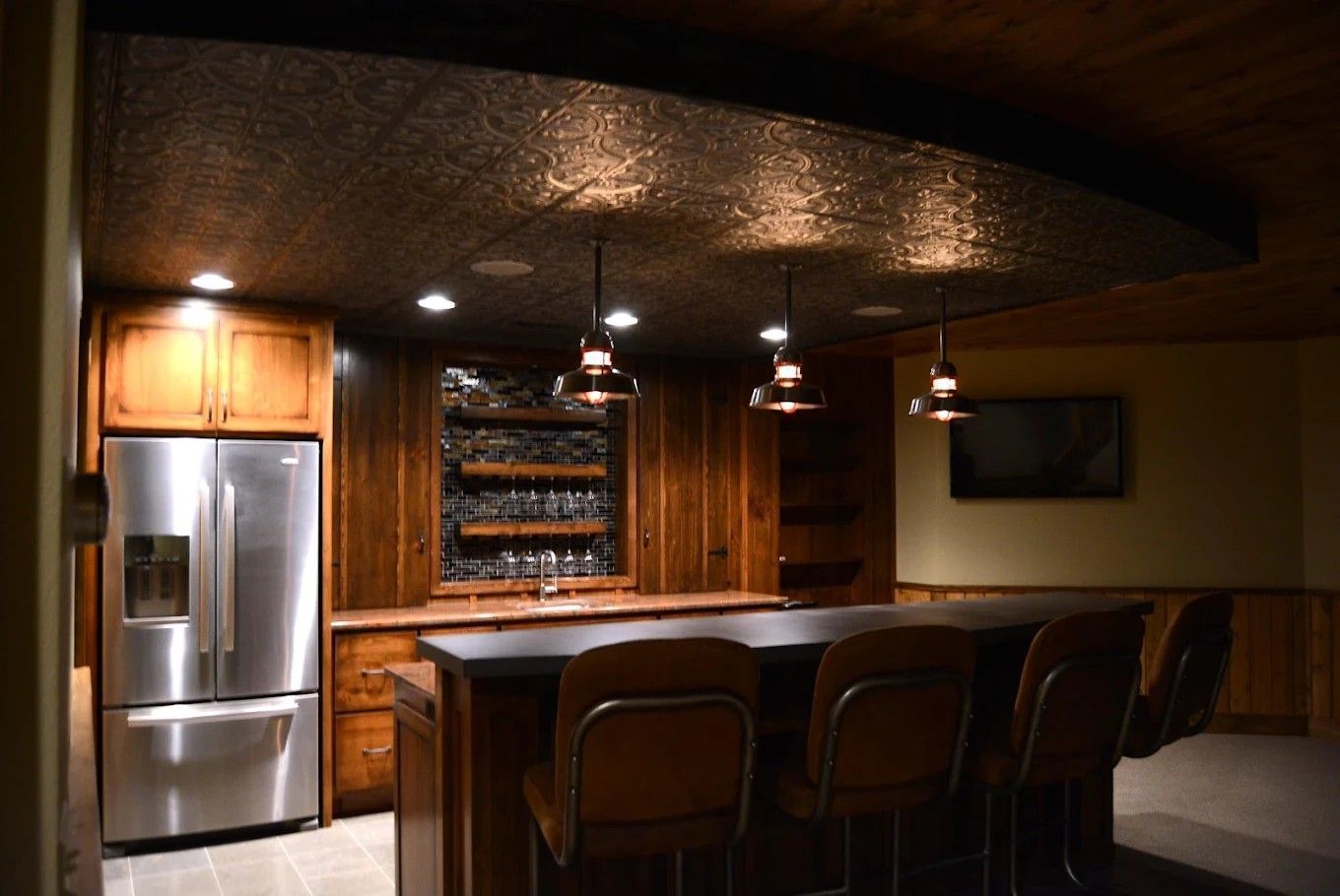 Modern kitchen with stainless steel fridge, wood bar counter, and pendant lights under a dark ceiling
