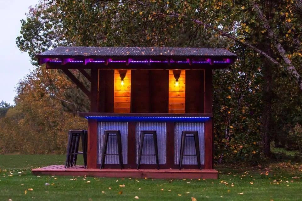 Small rustic outdoor cabin lit warmly at dusk, with purple lights and trees around it