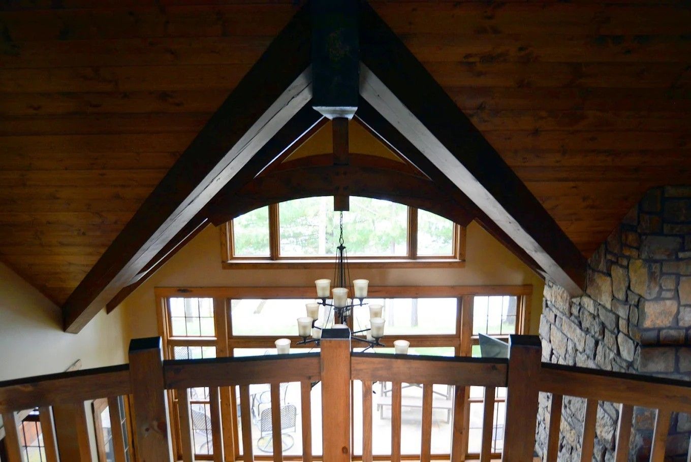 Wood-paneled loft with vaulted beams, stone wall, and a window overlooking a railing and chandelier