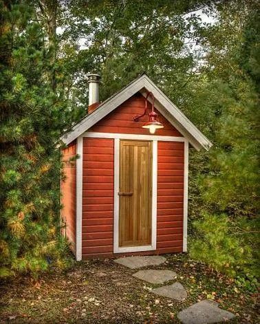 Small red wooden cabin with a light wood door, set among trees on a stone path.