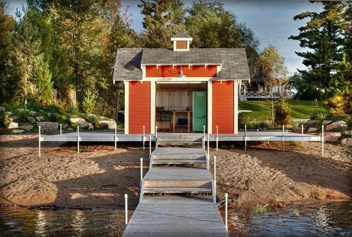 Small red lakeside cabin with dock and ramp, surrounded by trees in autumn.