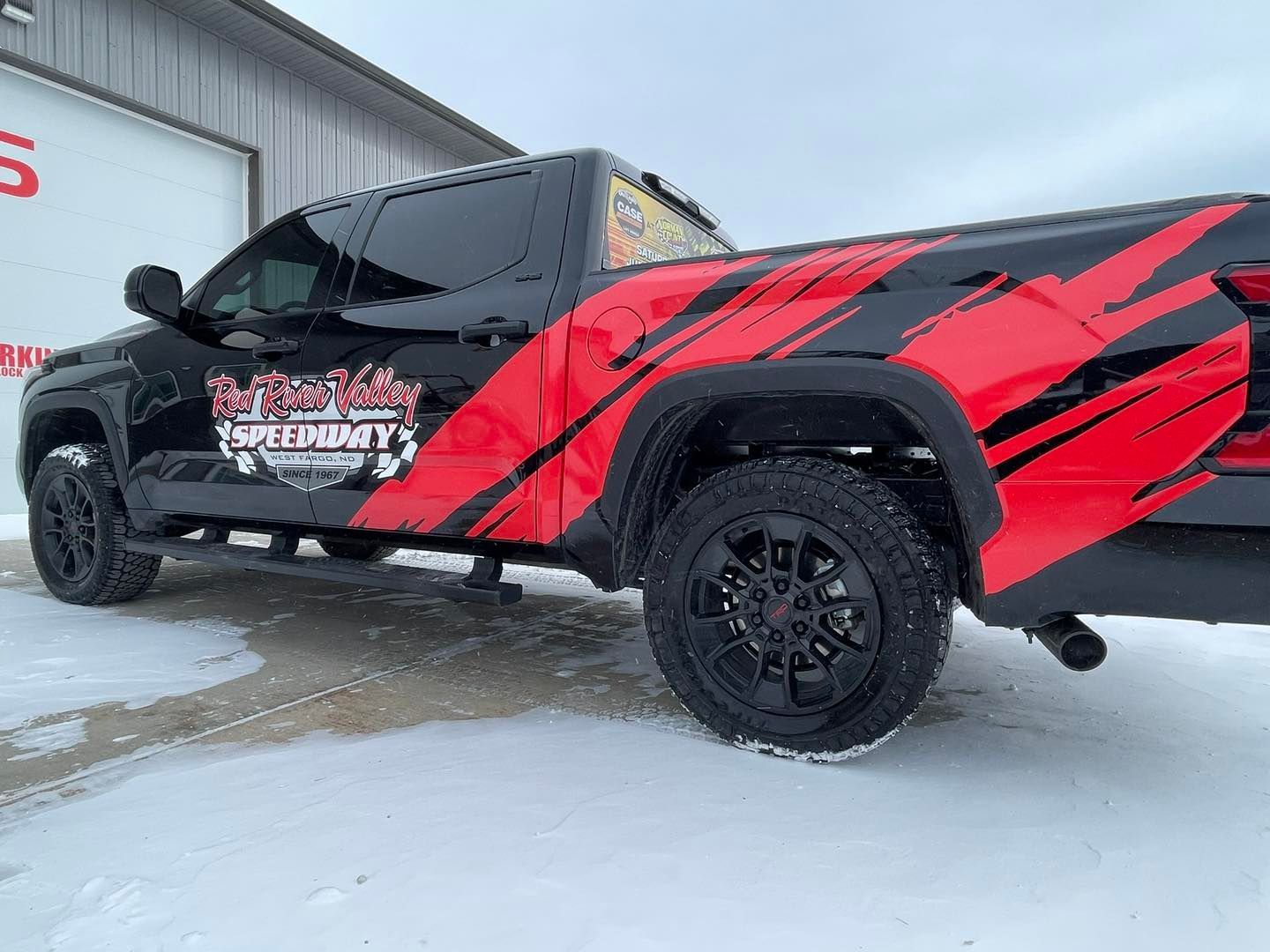 A red and black truck is parked in the snow in front of a building.