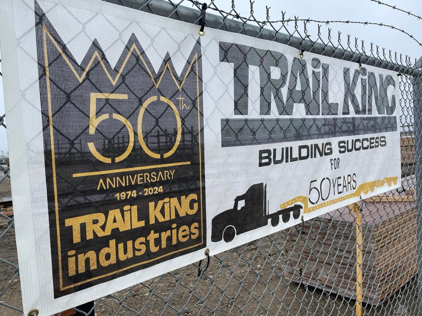 A banner for trail king industries is hanging on a chain link fence.