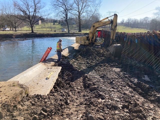 A man is standing next to a large excavator in the mud near a river.