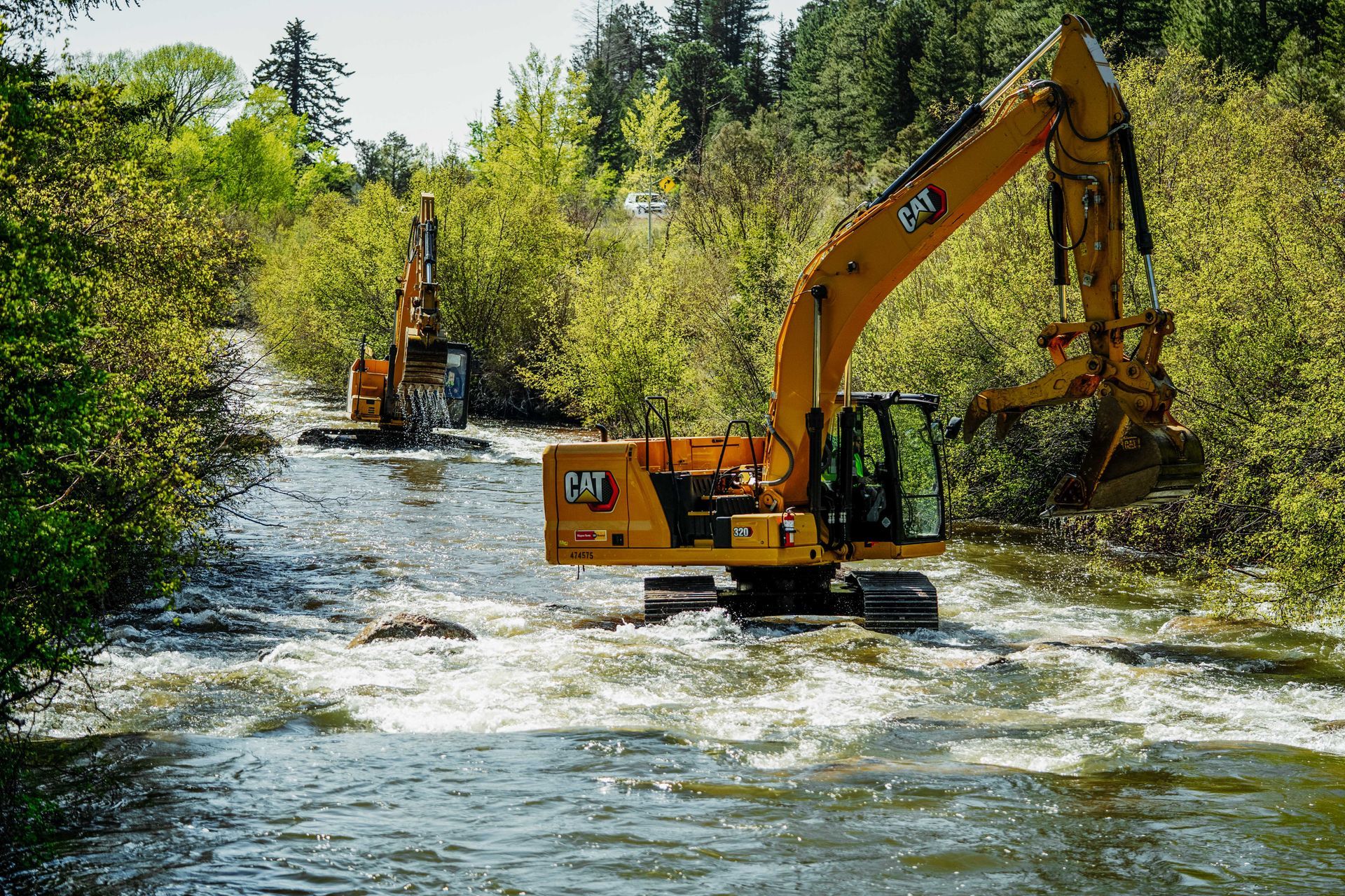 A yellow excavator is driving through a river surrounded by trees.