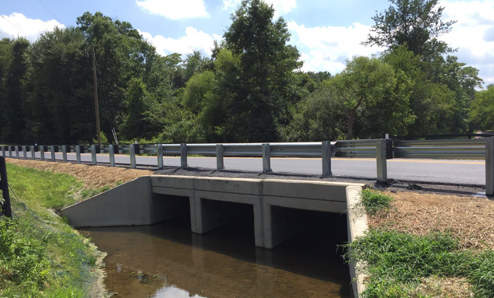 A bridge over a river next to a road with trees in the background.