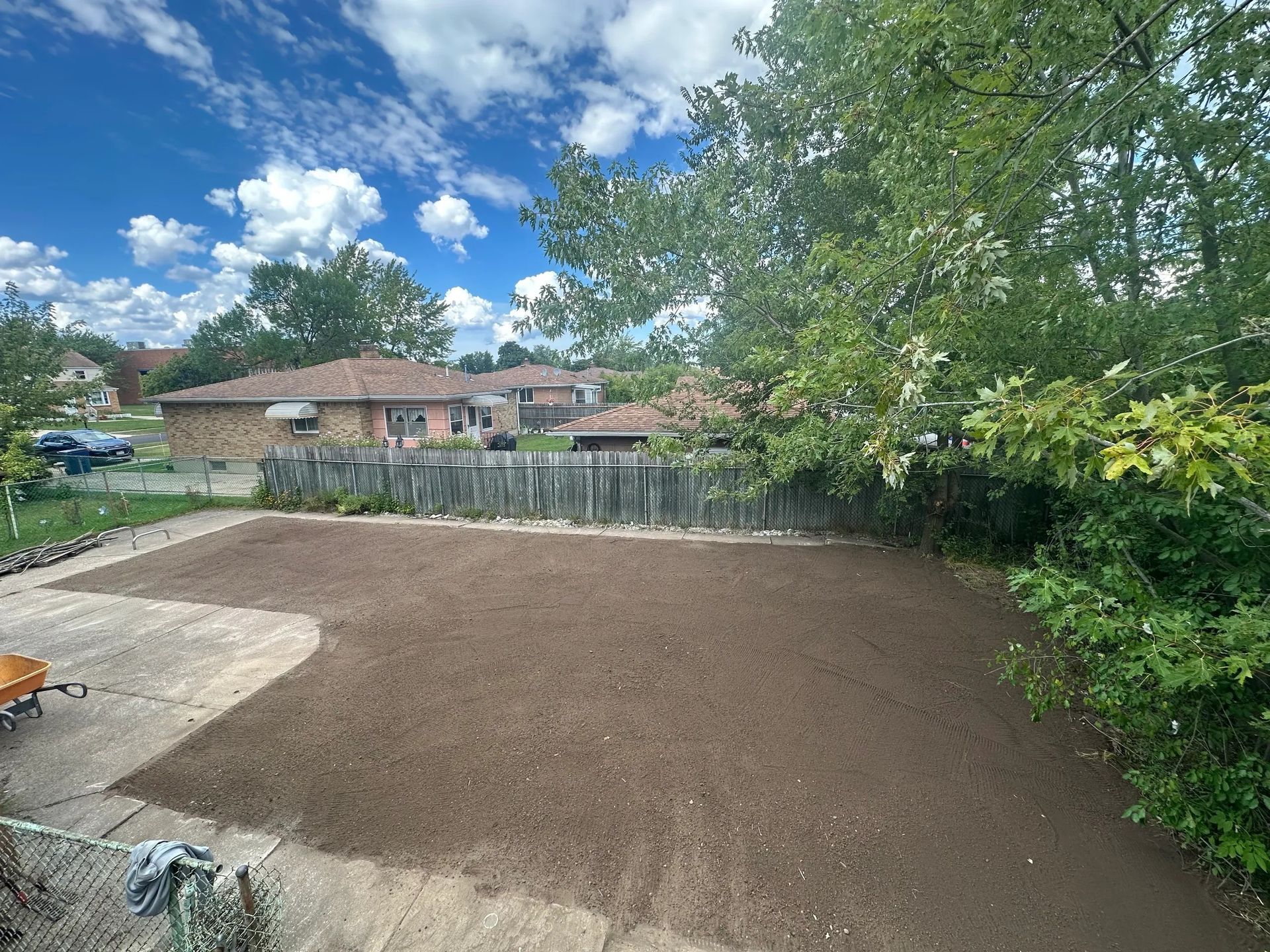 Freshly graded dirt lot beside a concrete patio, with trees, fence, and houses under a blue sky.
