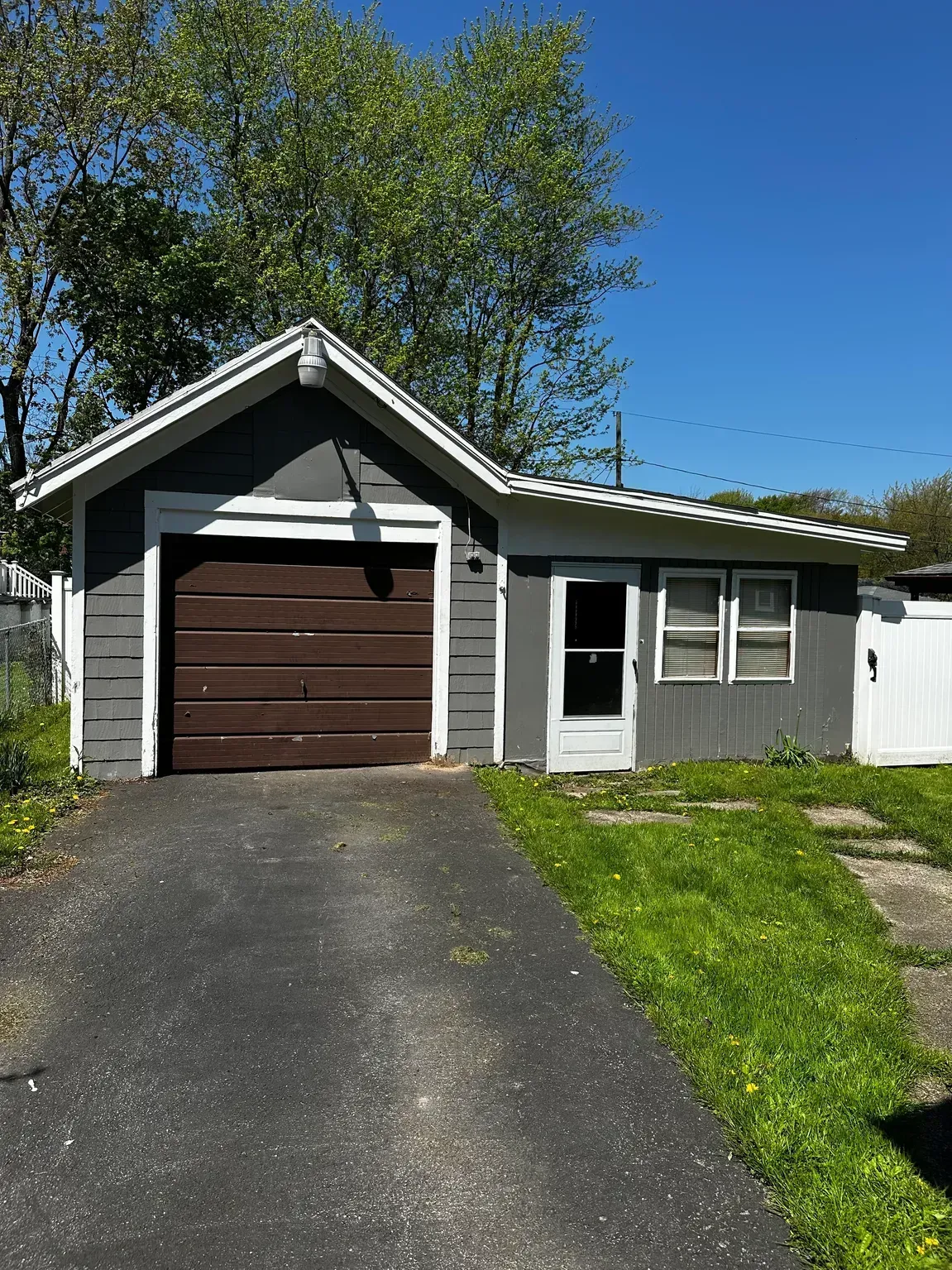 Small gray garage with a brown door and driveway, beside a white shed under a blue sky.