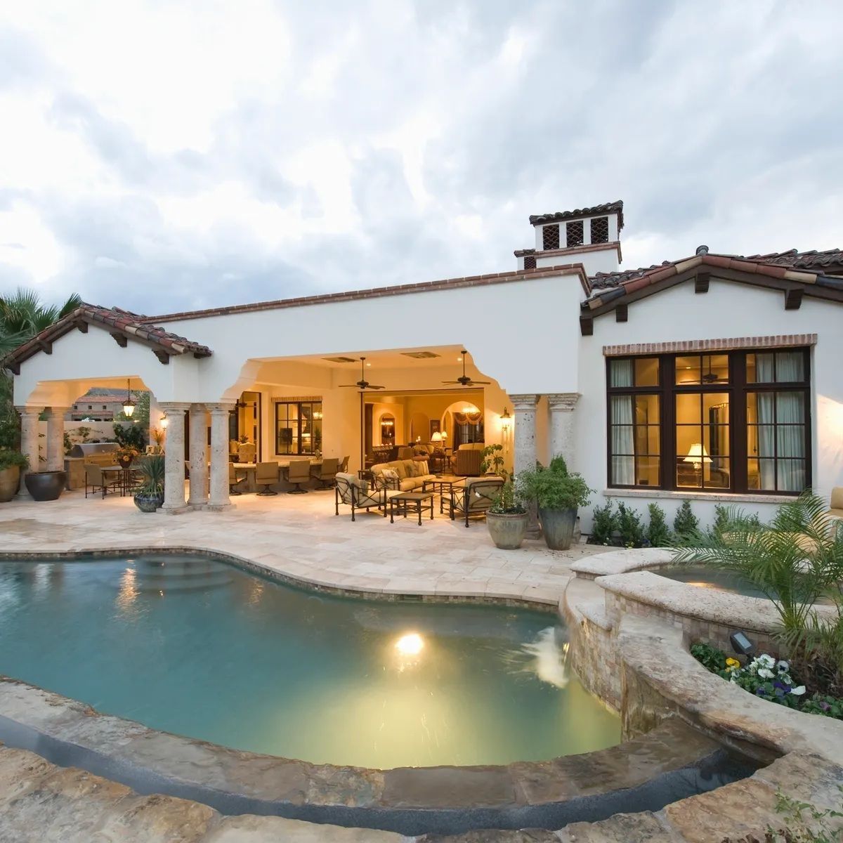 Spanish-style house with lit patio and curved backyard pool at dusk