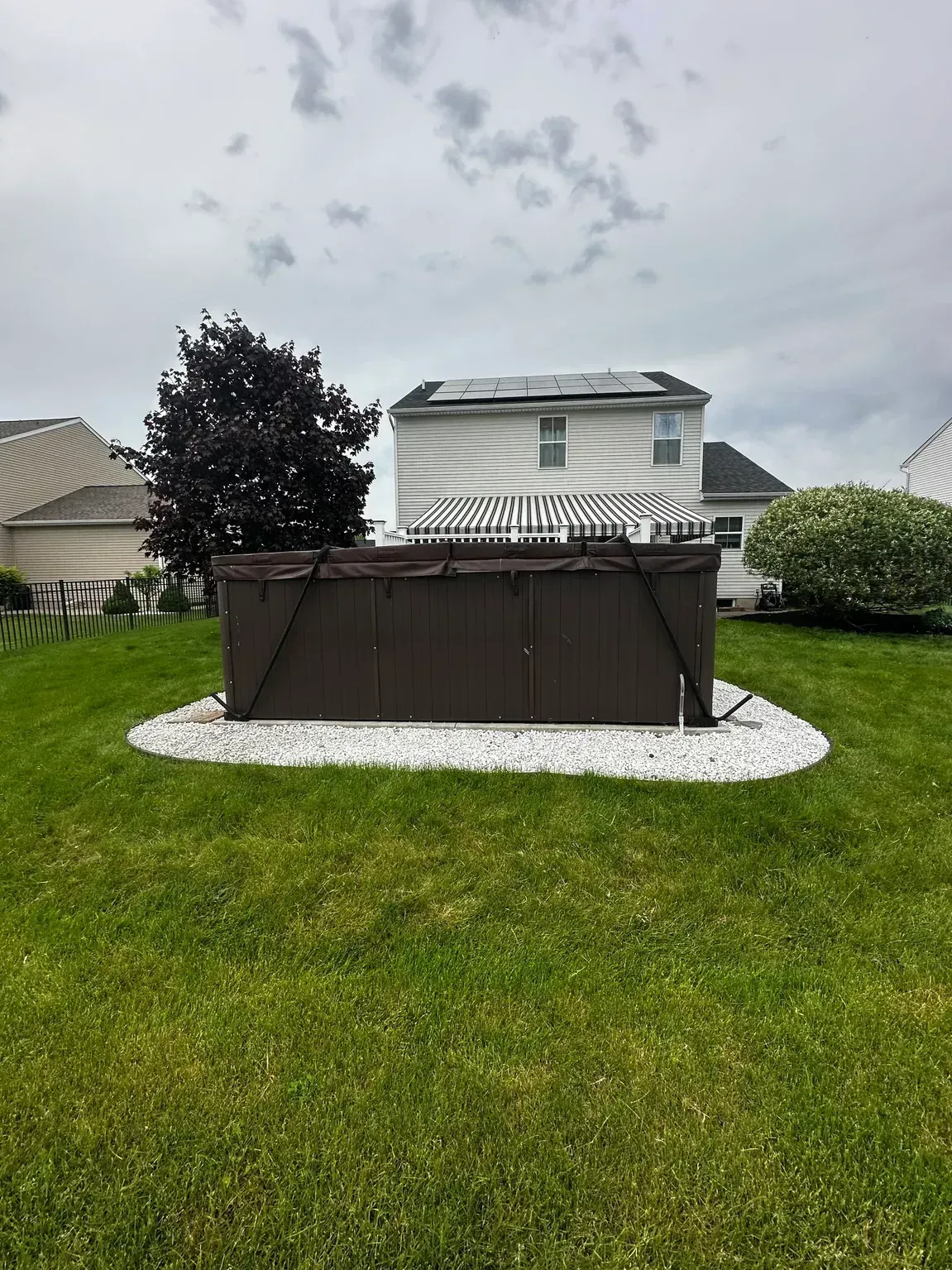Backyard with a brown hot tub on a white gravel pad, surrounded by green grass and houses under a cloudy sky