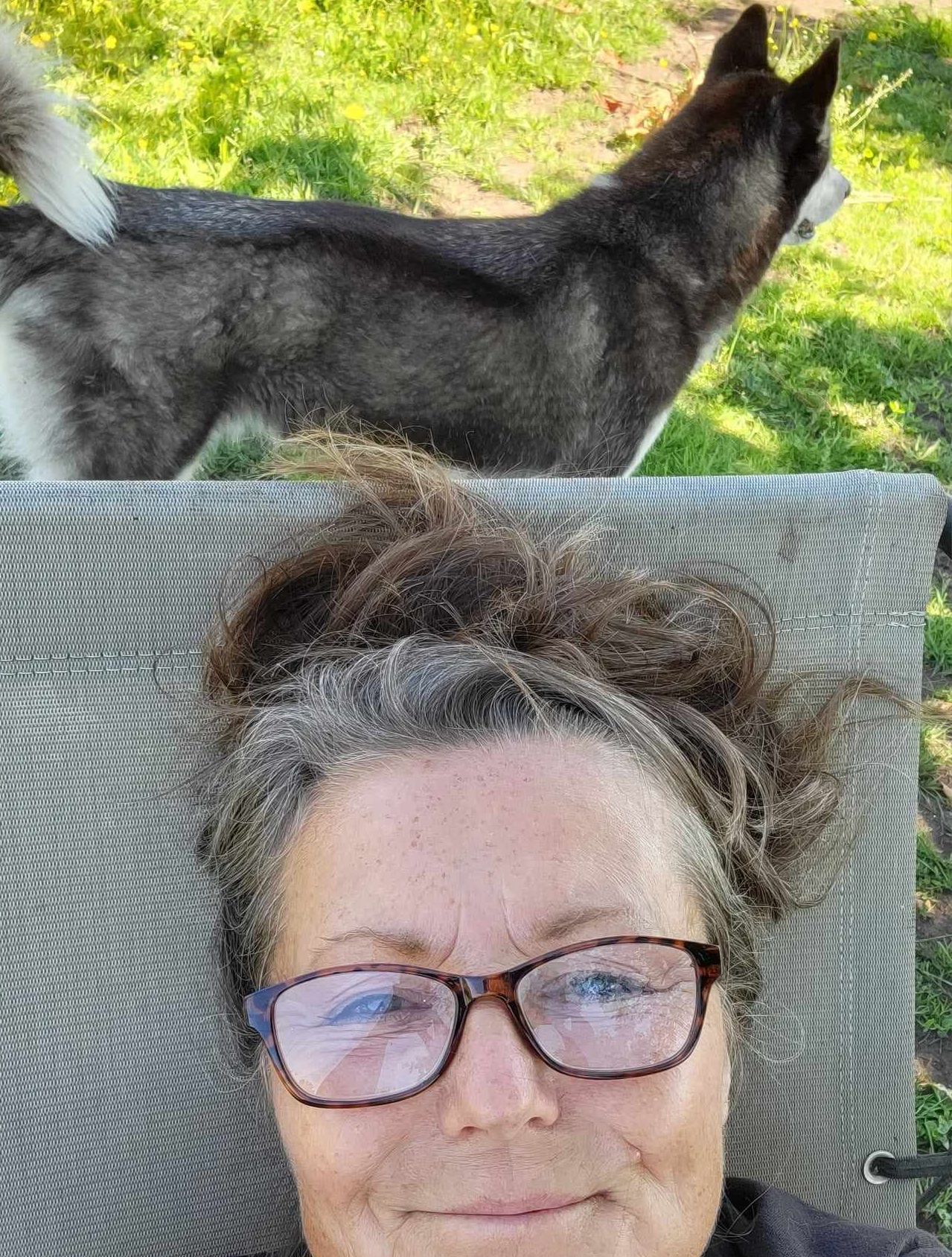 A woman wearing glasses is laying in a chair with a dog behind her.