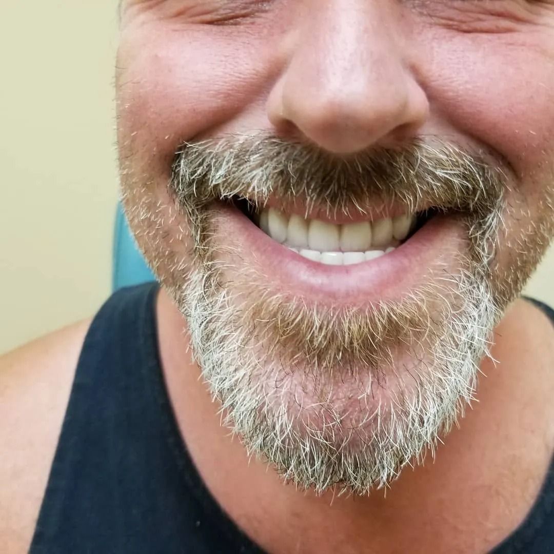 Man with a gray beard and smiling, showing white teeth. He wears a black tank top.