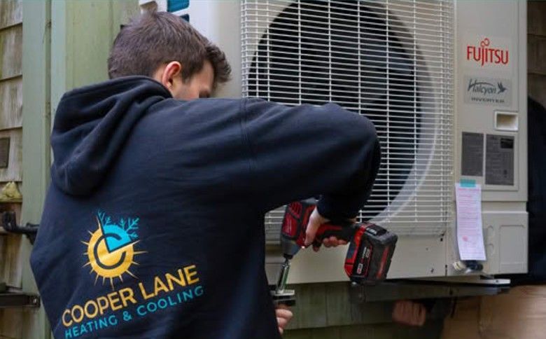 HVAC technician in black hoodie using a power drill on a Fujitsu air conditioning unit mounted on a wooden wall.