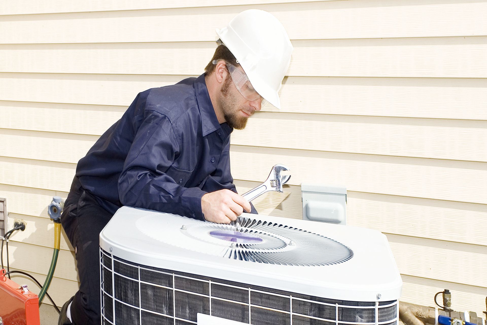A technician in a hard hat inspects an outdoor air conditioning unit next to a light-colored building.