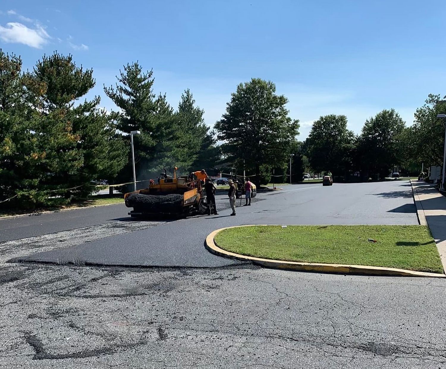 Road crew with orange equipment paving a curved asphalt lot beside trees and a grassy median under a clear sky