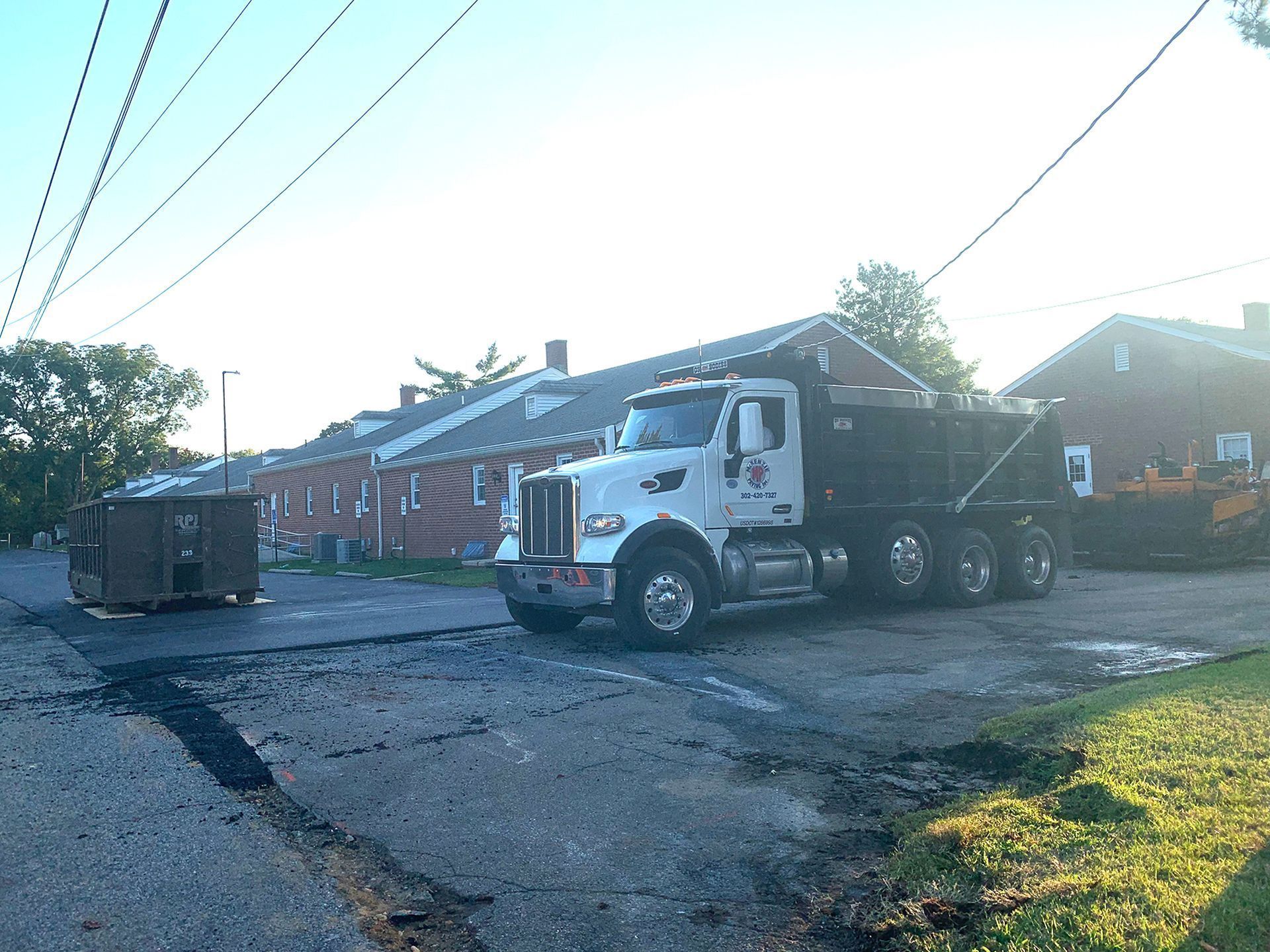 White dump truck on a rough street beside brick houses and a dumpster in bright sunlight.