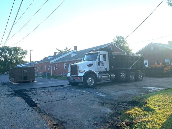 White dump truck on a rough street beside brick houses and a dumpster in bright sunlight.