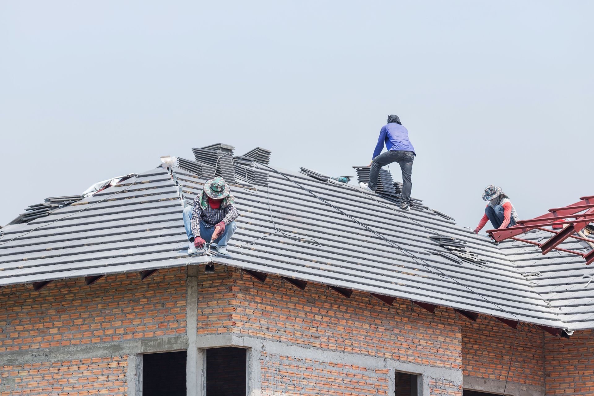 Workers repairing a rooftop on a brick building, stacking gray shingles under a cloudy sky