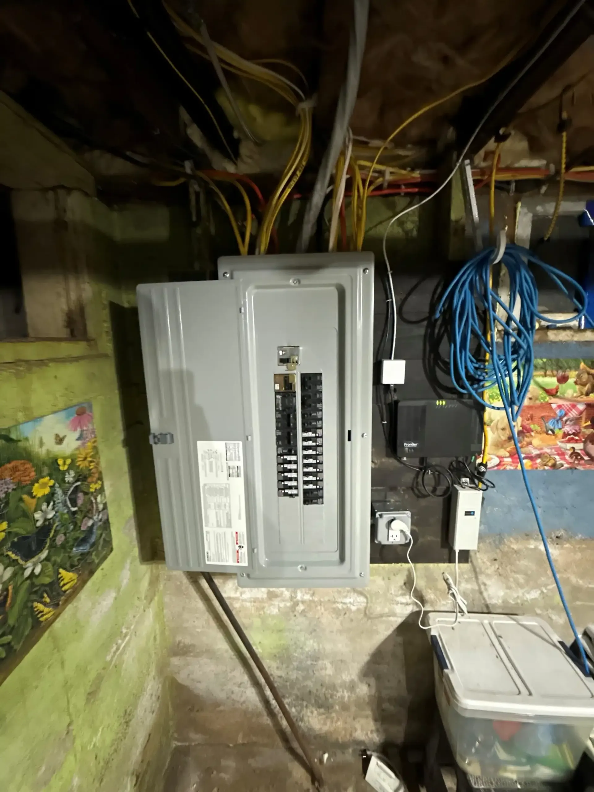 Electrical panel with open door in a basement, surrounded by wires, with a colorful mural and utility box.