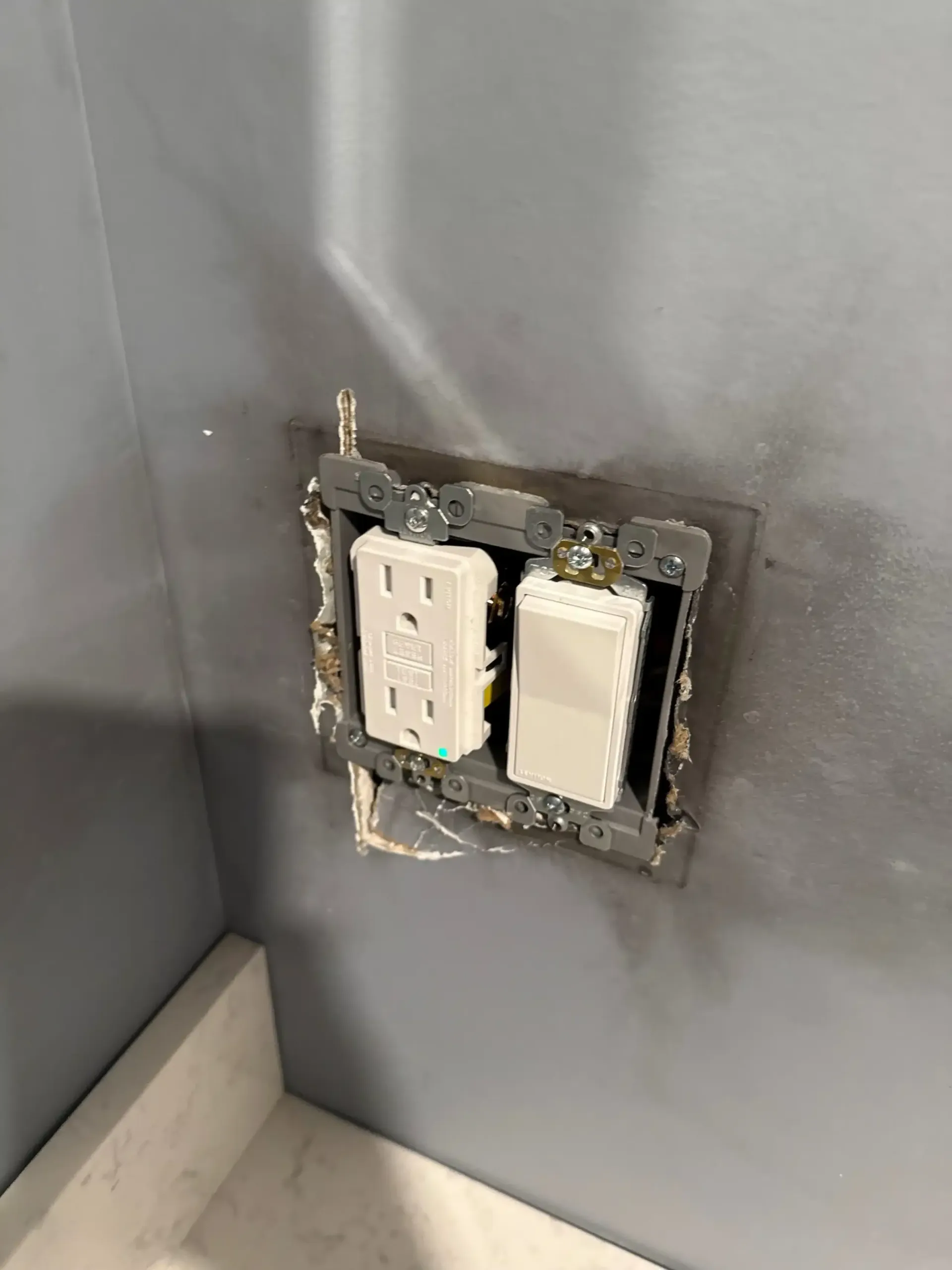 Gray wall with an exposed electrical outlet and switch.