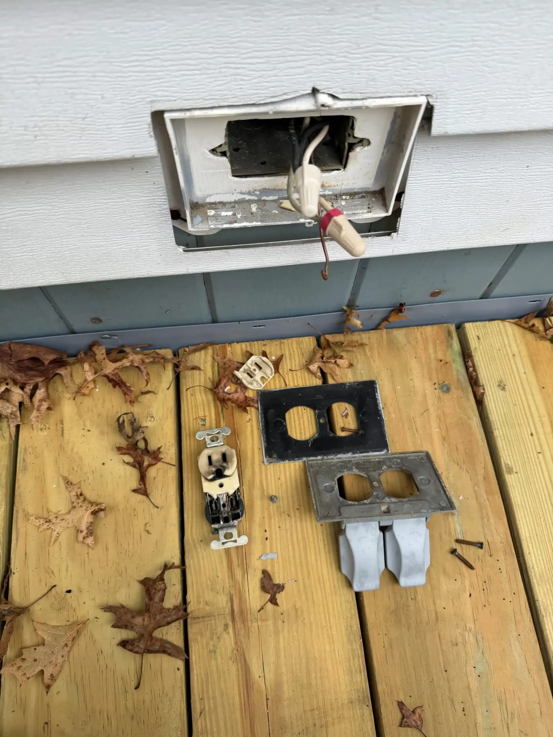 Outdoor electrical outlet parts scattered on wooden decking. Gray siding background.