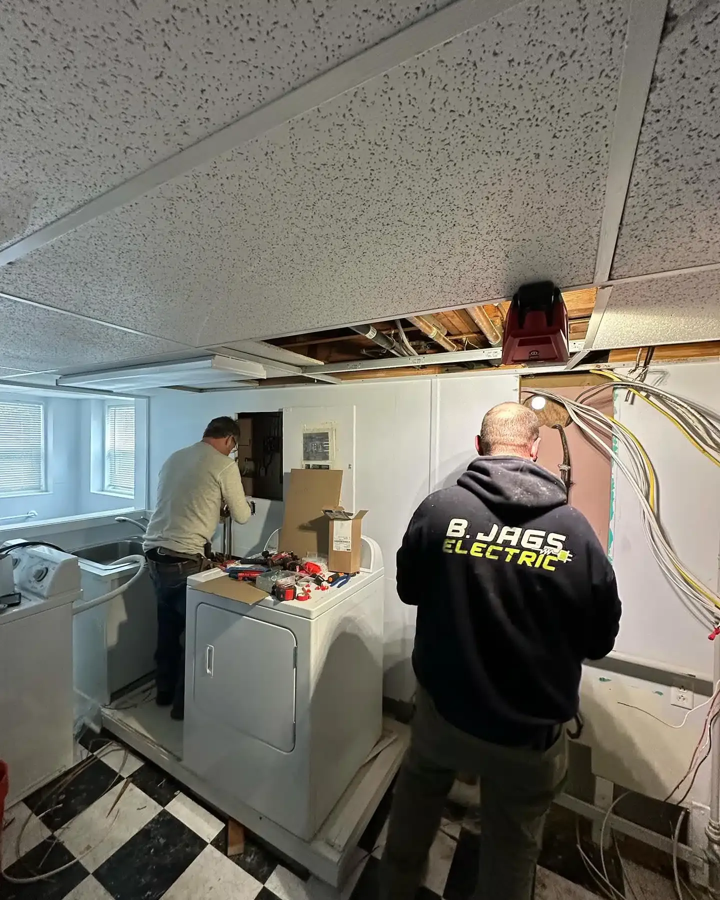 Two electricians work on a panel above a washer and dryer in a basement with a damaged ceiling.