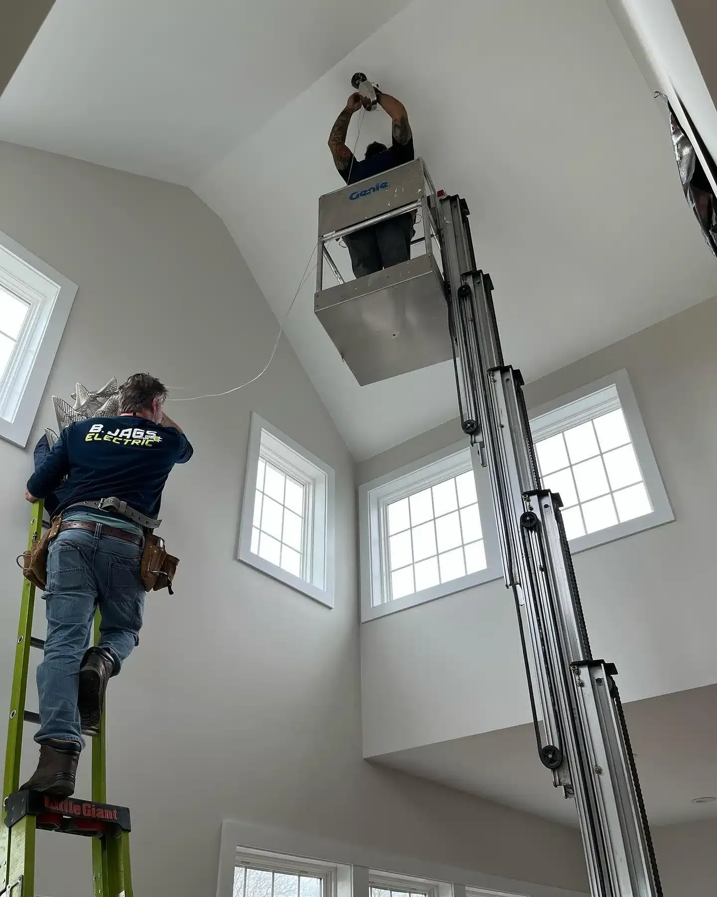 Two workers installing lights in a tall room, one on a lift, one on a ladder.