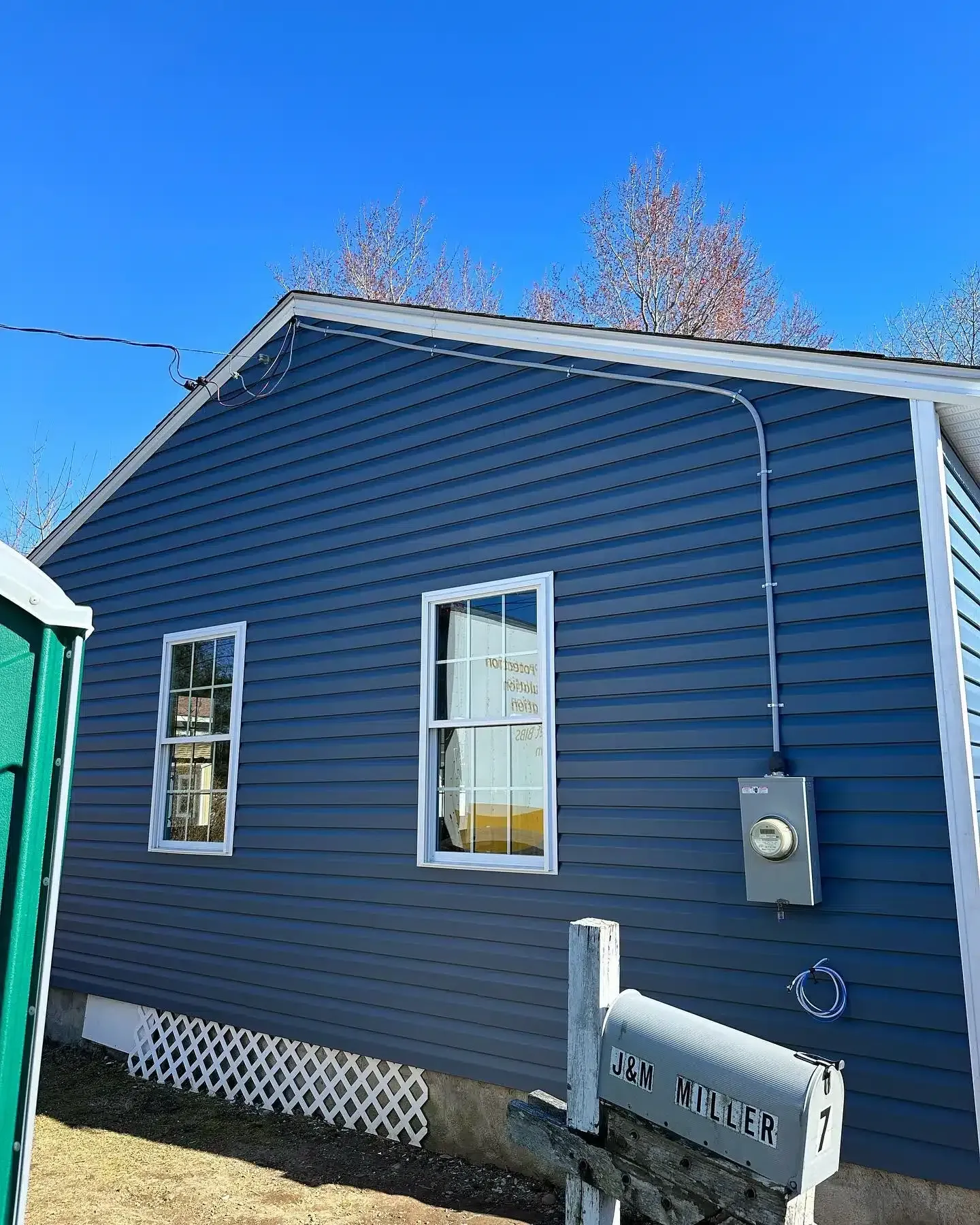Blue-sided house with white-framed windows, mailbox, and electrical box under a clear blue sky.