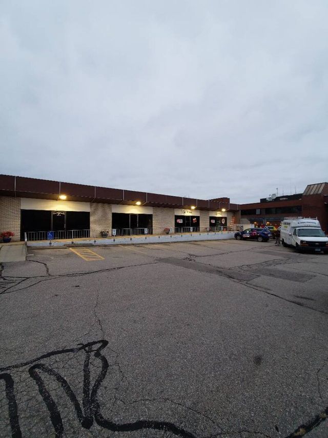 Exterior view of a commercial building with multiple storefronts and a cracked asphalt parking area under a cloudy sky.