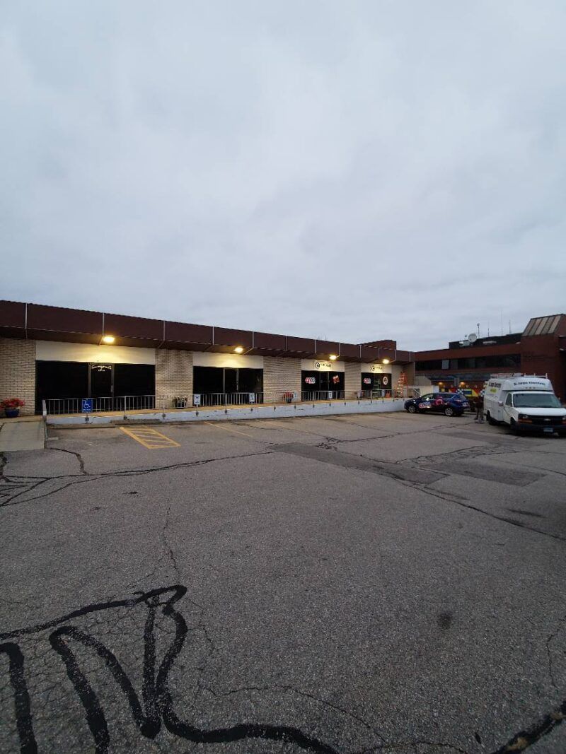 Exterior view of a commercial building with multiple storefronts and a parking lot under an overcast sky.