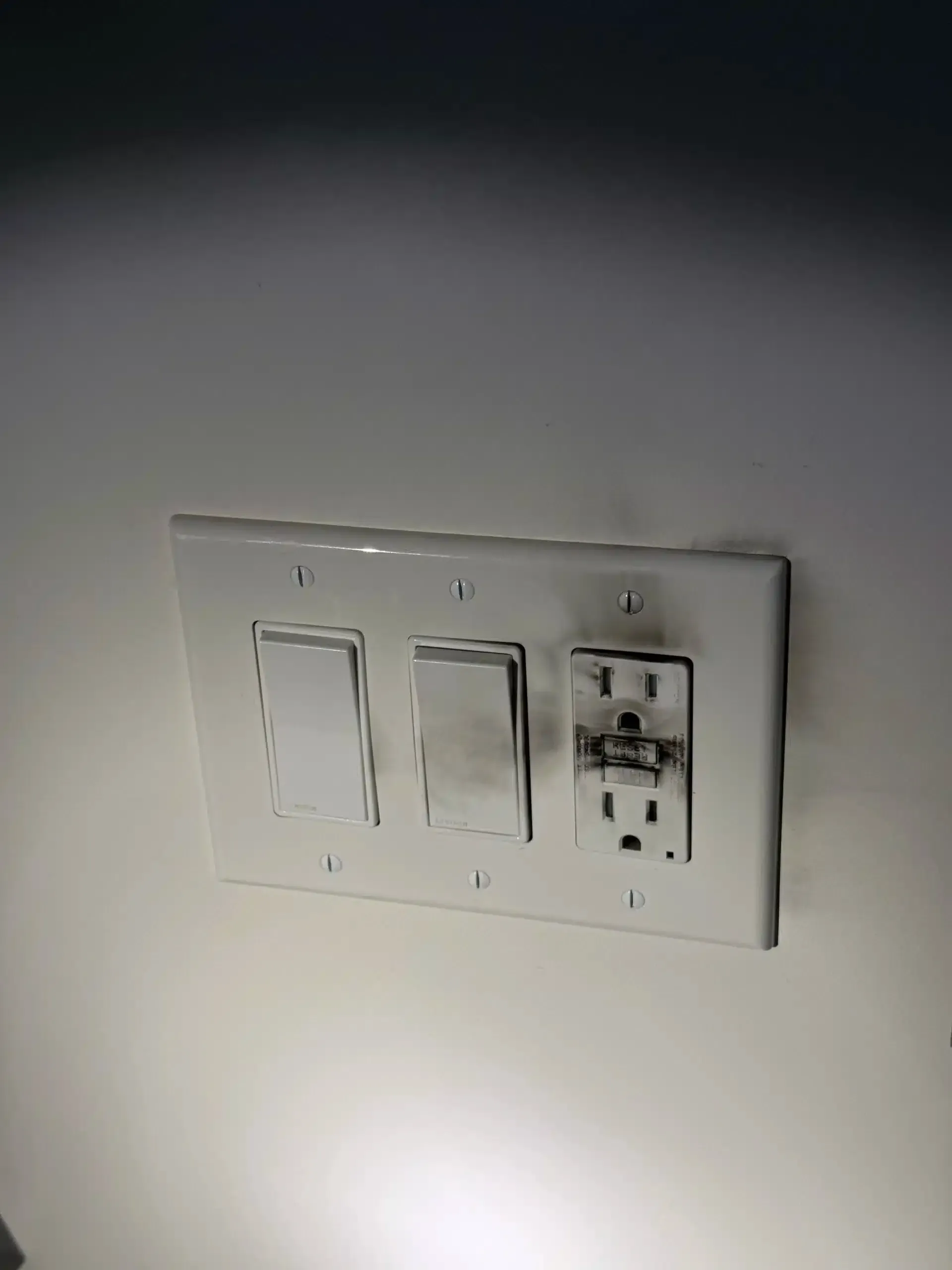 White electrical switch and outlet plate on a white wall, with burn marks on the middle switch and outlet.