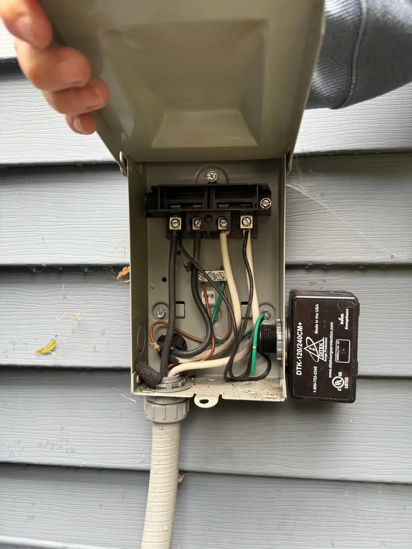 Open electrical box with wiring against gray siding. A hand holds the lid.