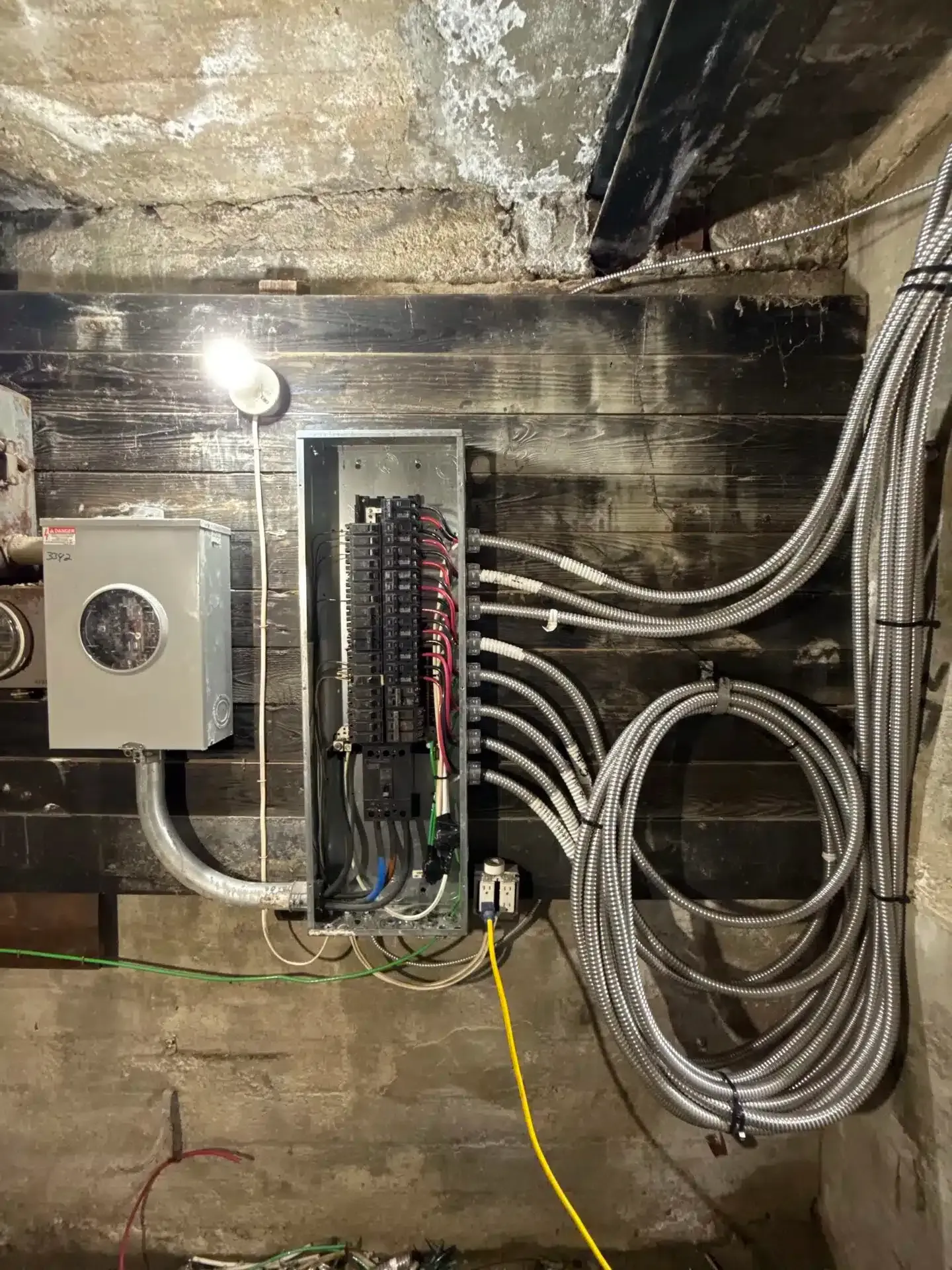 Electrical panel with wiring on a dark wood and concrete wall. A lightbulb illuminates the panel.