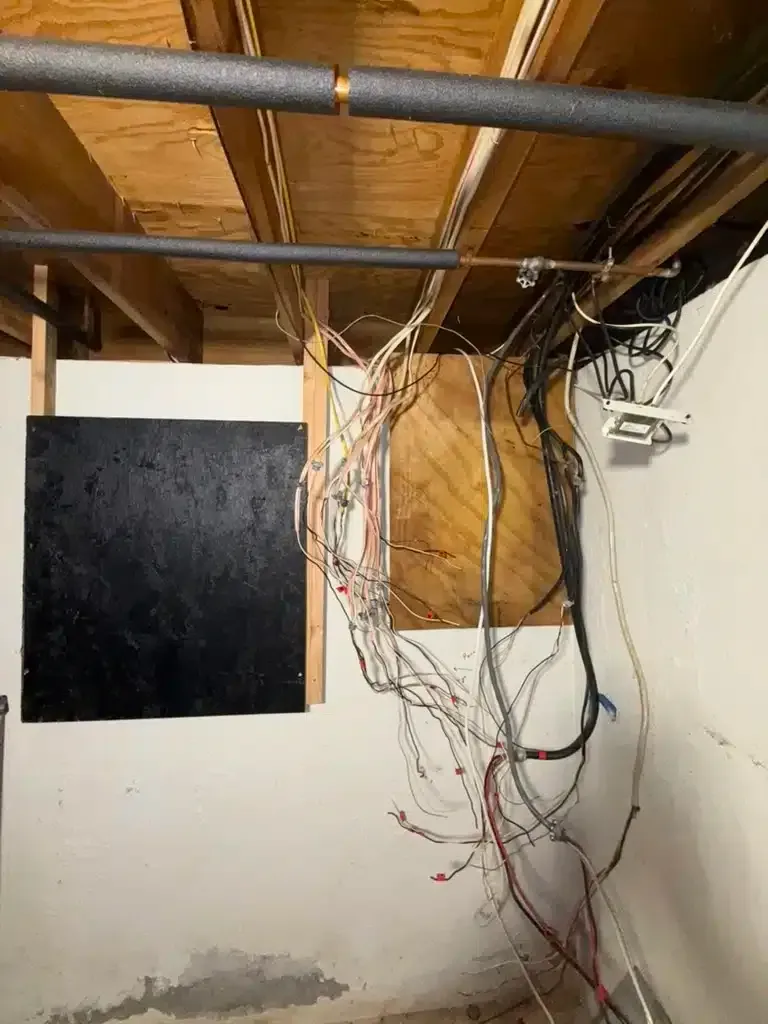 Exposed electrical wiring hanging from a wooden panel in a basement; white wall, wooden ceiling, pipe.